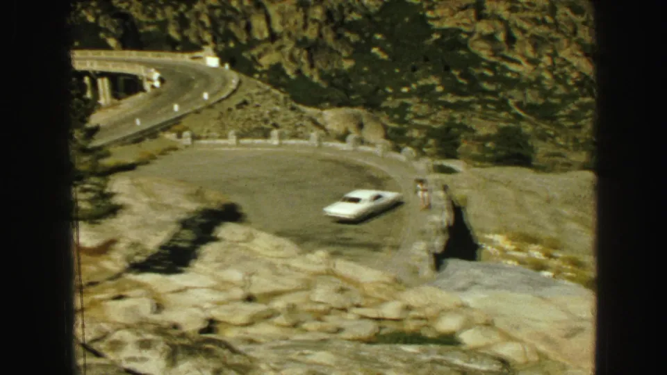 1968 Vancouver-area 8mm frame showing a white car rounding a mountain road curve while a person stands near the roadside edge.