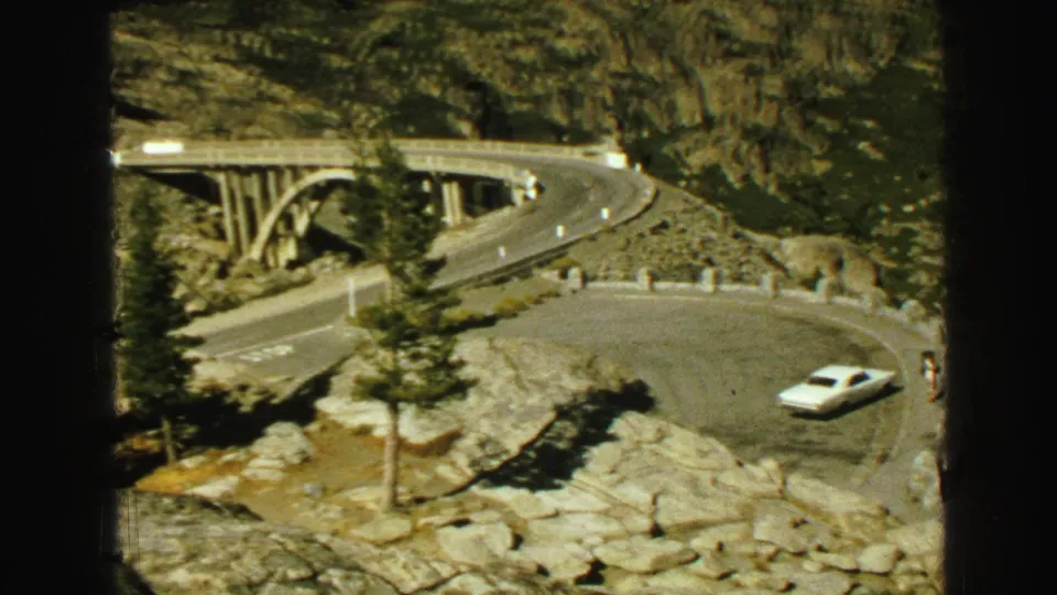 1968 Vancouver-area home-movie frame with a white car on a curved mountain road and an arched bridge visible beyond the trees.