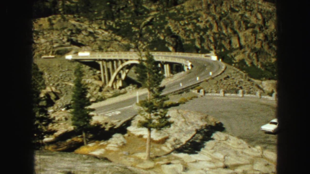 1968 8mm Vancouver-area travel frame showing a concrete arch bridge, winding road, rocky terrain, pine trees, and a partial white car.