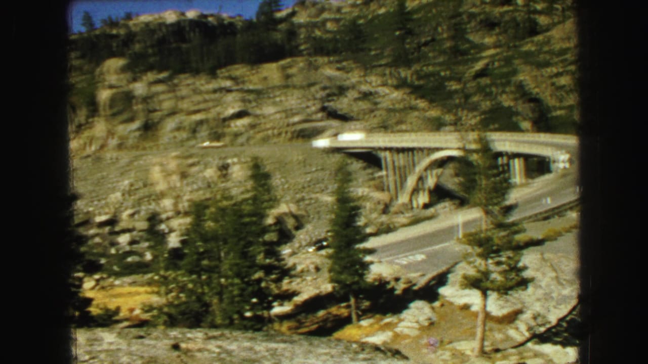 1968 8mm frame from Vancouver metadata showing a concrete bridge, road curve, pine trees, rock faces, and blue sky during a roadside pan.
