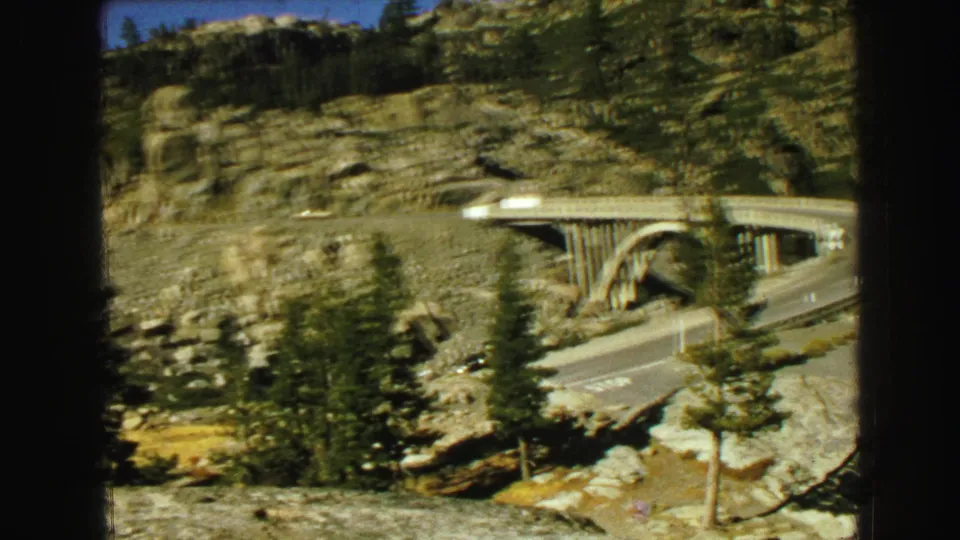 1968 8mm frame from Vancouver metadata showing a concrete bridge, road curve, pine trees, rock faces, and blue sky during a roadside pan.