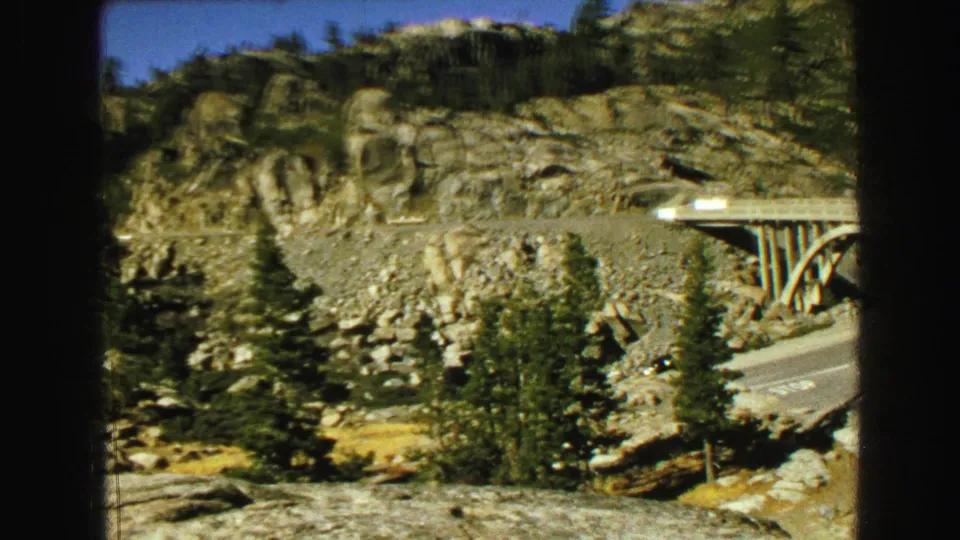 1968 8mm Vancouver-area frame with the bridge partly visible at right, a road below, evergreen trees, and sunlit rock formations.
