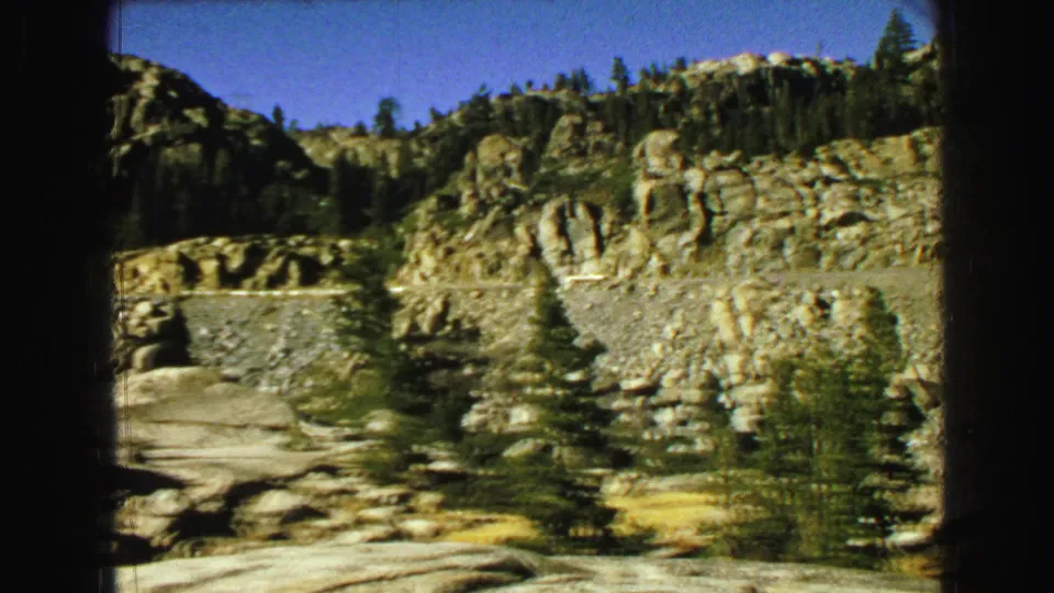 1968 8mm travel frame showing rugged rock formations, a winding road, evergreens, and the edge of a bridge in the Vancouver metadata location.