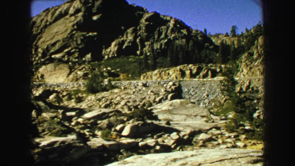 Vintage 8mm frame showing a rugged rock outcrop, roadside stonework, and evergreens during a Vancouver-area mountain drive.