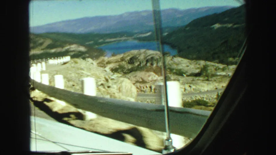 1968 car-window view of a lake, distant hills, roadside barriers, and windshield frame in British Columbia mountain scenery.