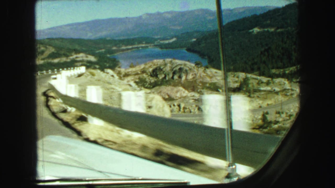 Car-window view of a lake, white roadside barriers, rocky slope, and distant mountains.