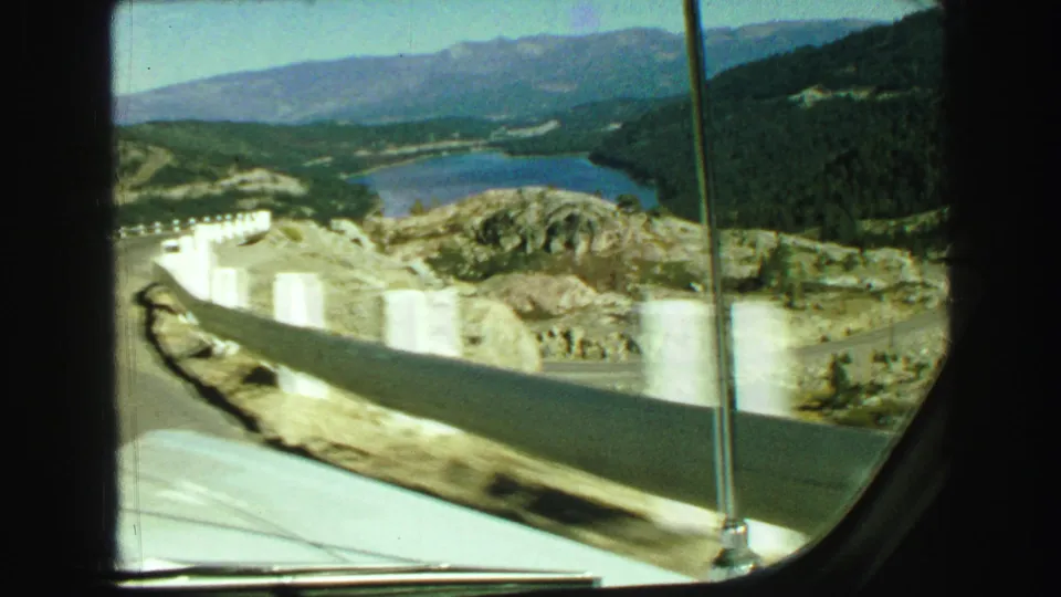 1968 Vancouver-area 8mm car-window view showing a lake, pale roadside barriers, rocky terrain, wooded hills, and mountains beyond.