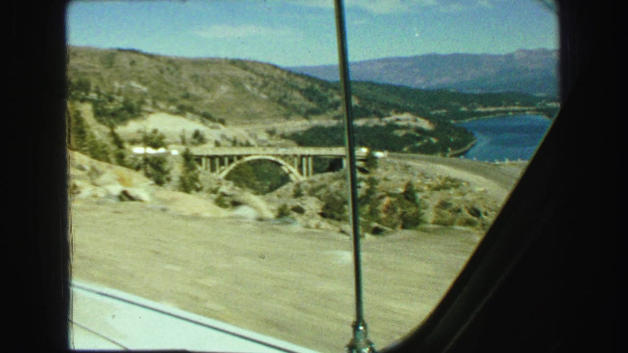 Arched bridge crossing a rocky valley near a lake and forested hills.