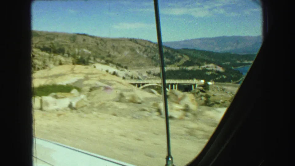 1968 Vancouver-area 8mm frame showing motion-blurred rocky roadside ground, an arched bridge, lake water, and hills through the car window.