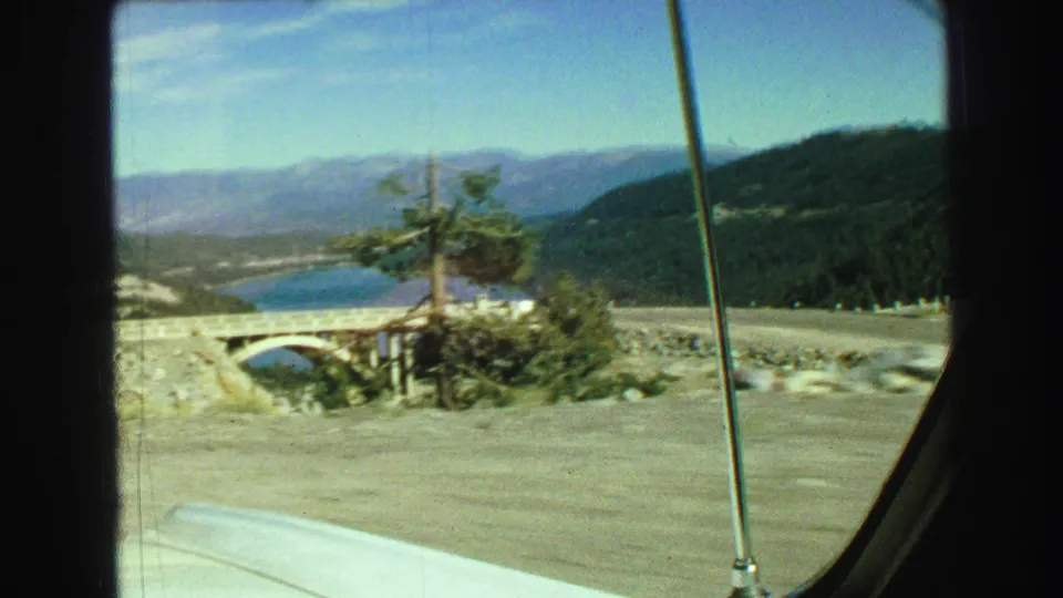 1968 Vancouver-area travel frame with a lake, distant mountains, a bridge, roadside shrubs, and the edge of the car window in view.