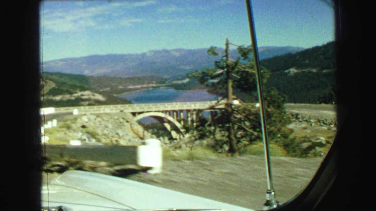 1968 8mm car POV near Vancouver, British Columbia, showing a road, bridge, lake, forested hills, and distant mountains.