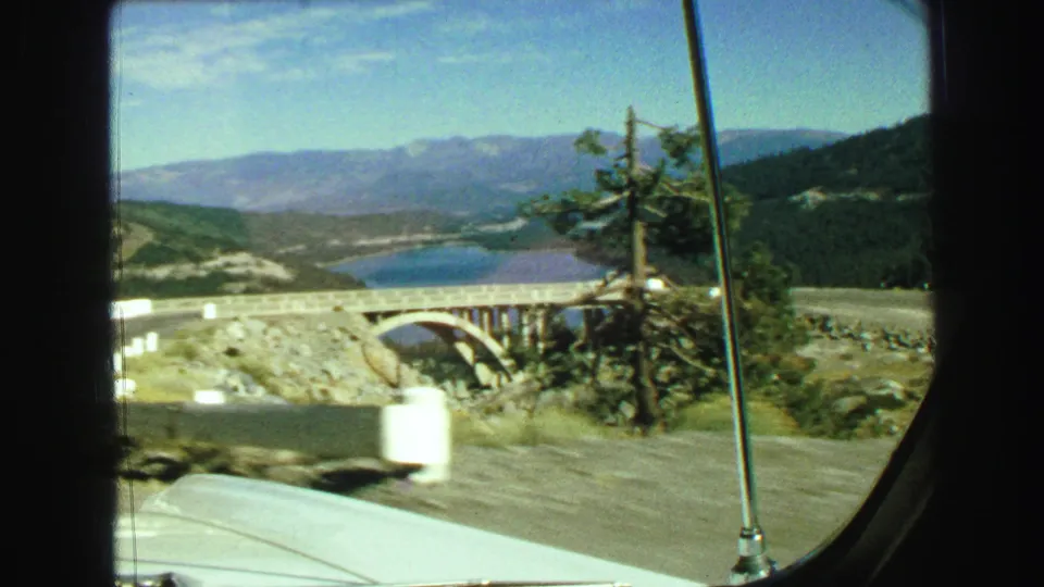 1968 8mm car POV near Vancouver, British Columbia, showing a road, bridge, lake, forested hills, and distant mountains.