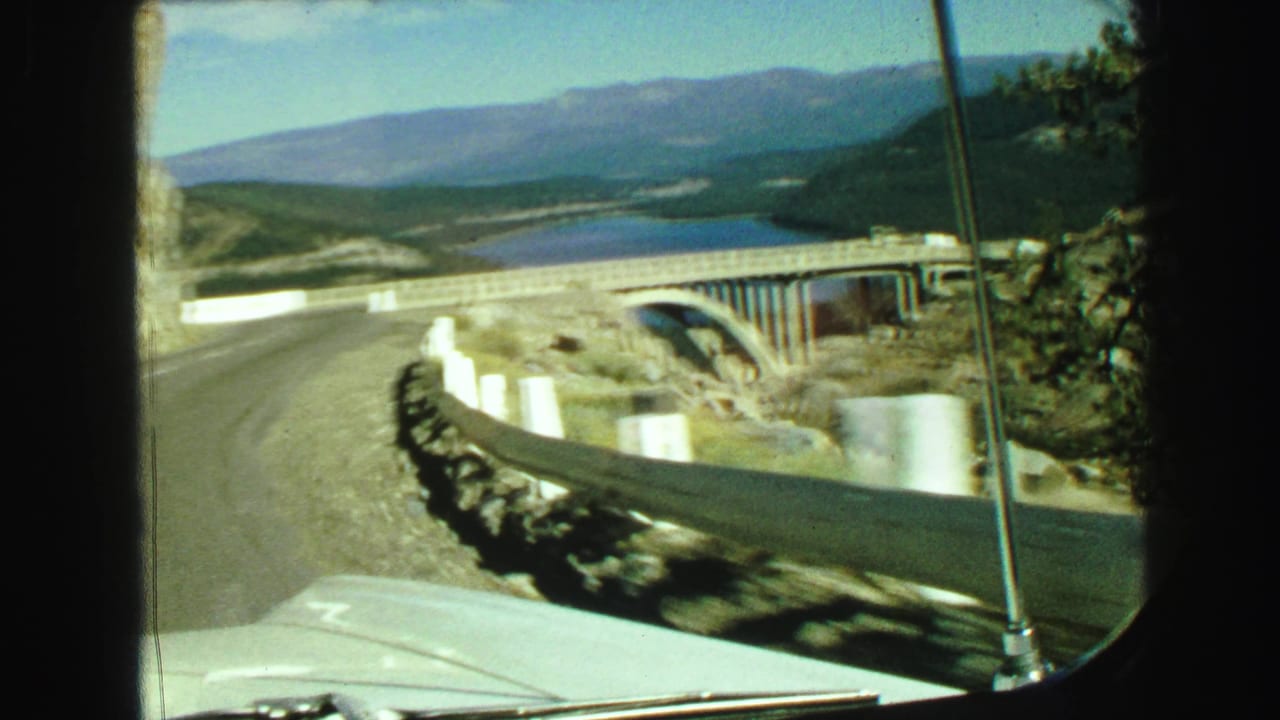Road curves toward a concrete bridge with lake water and mountains beyond the windshield.