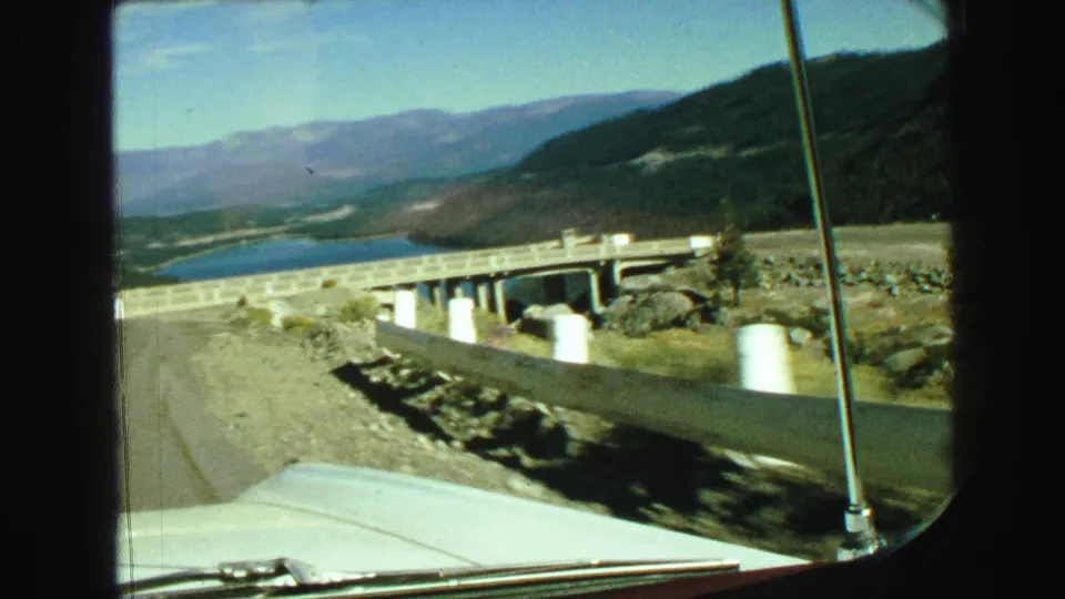Vintage 8mm car view in British Columbia showing a roadside guardrail, bridge structure, lake, hills, and pale car hood.