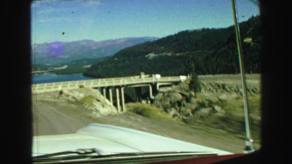 1968 Vancouver-area car POV looking past the hood and antenna toward a bridge, lake, forested slope, and distant peaks.