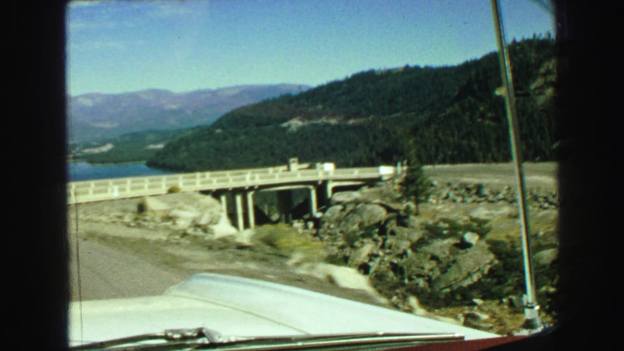 Car hood and windshield frame approaching a concrete bridge beside a lake and wooded hillside.