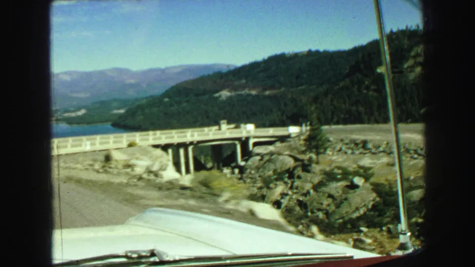 1968 8mm road-view frame from Vancouver-area metadata, showing a car approaching a concrete bridge with water, rocks, forested hills, and distant mountains.