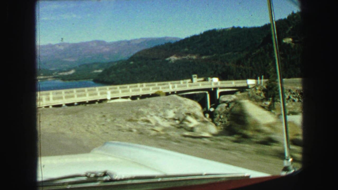 Car moves closer to a concrete bridge with rocks, water, forested hills, and mountains beyond.