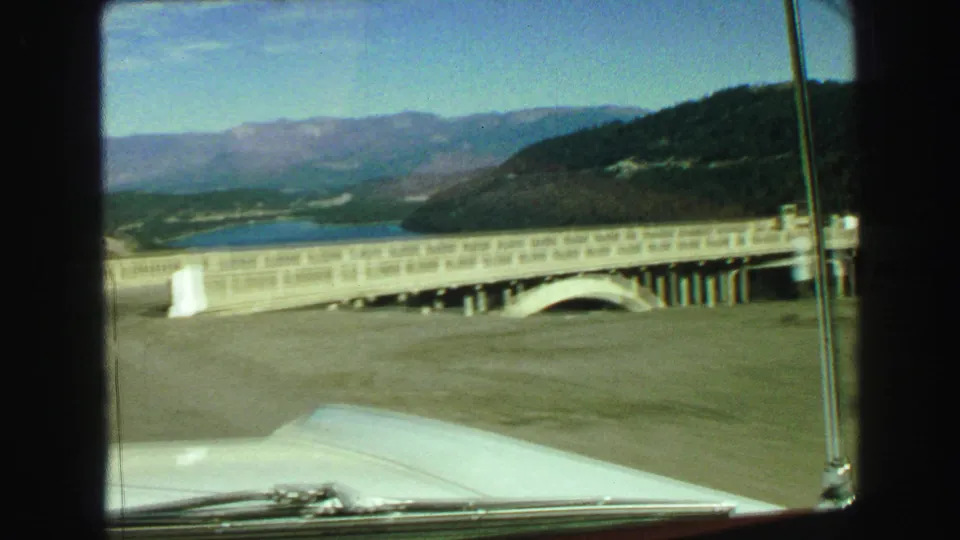 Inside-car view during a 1968 Vancouver-area bridge crossing, showing the concrete rail, car hood, windshield wiper, lake, hills, and distant peaks.