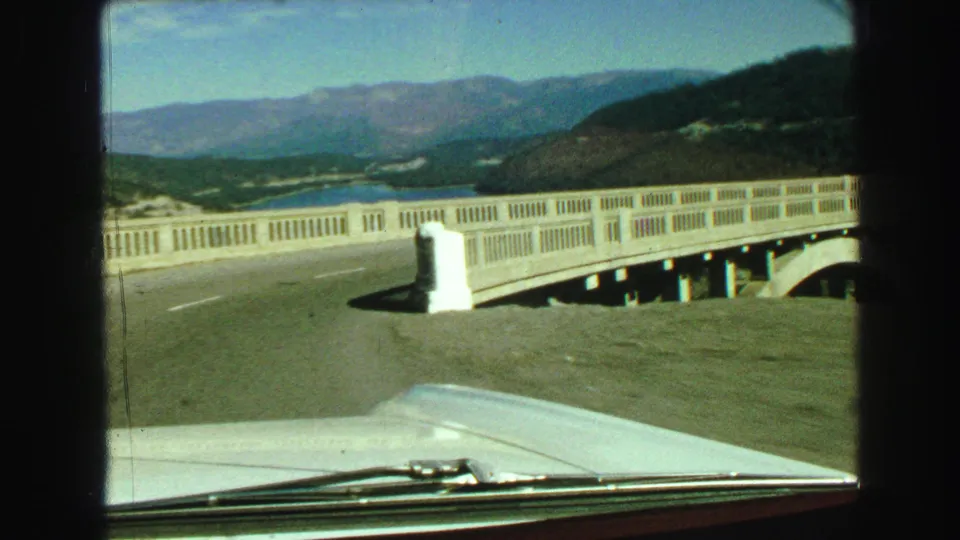 1968 8mm home-movie frame of a car traveling along a concrete bridge in British Columbia, with water, wooded hills, and mountains beyond.