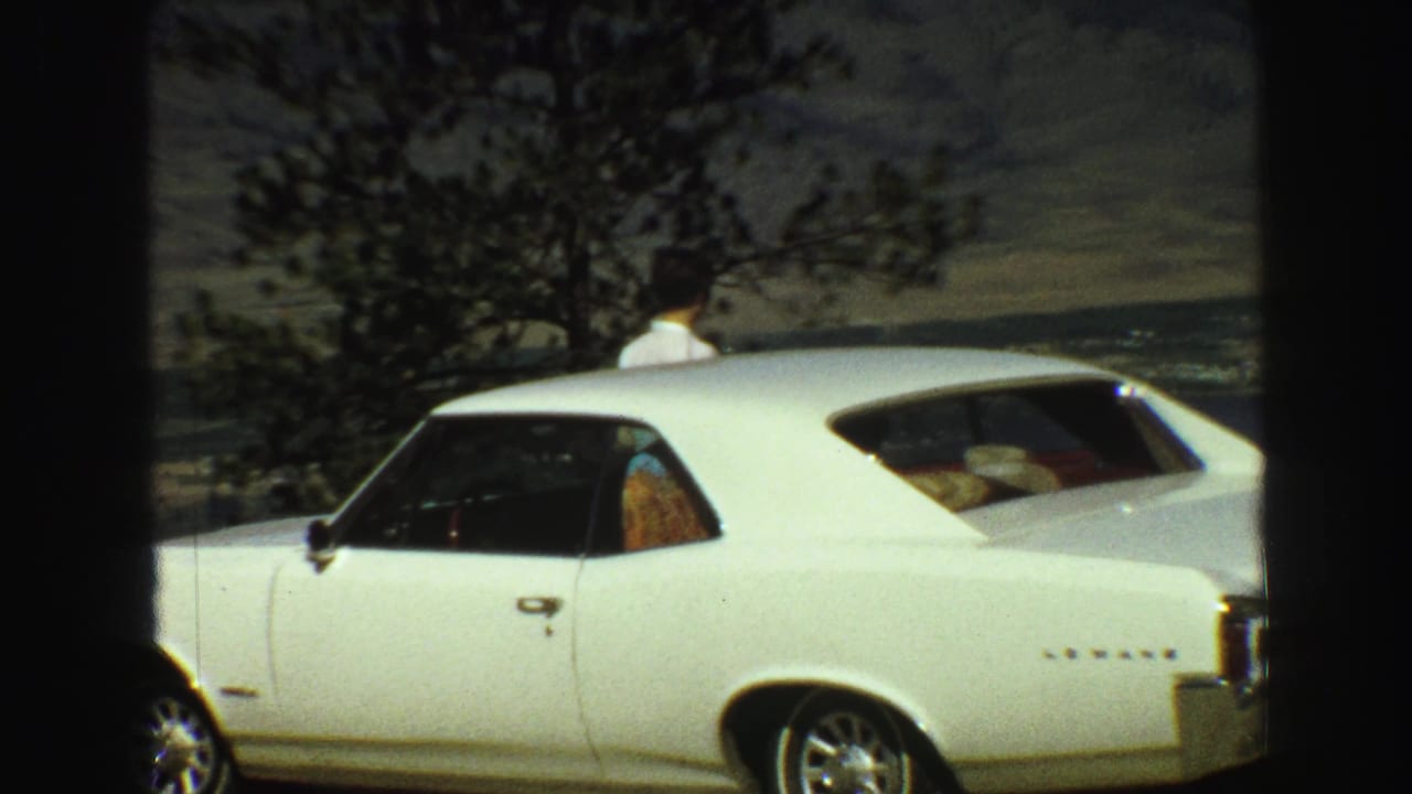 White two-door car beside a scenic overlook with a person partly visible behind it.