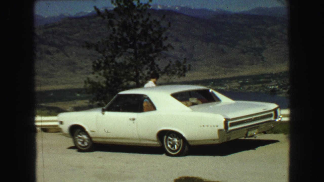 White car parked near a guardrail with mountains, water, and a person in the background.
