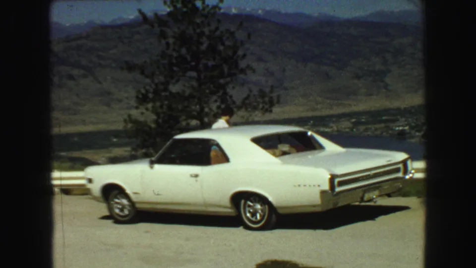 A white late-1960s-style car is parked at a Vancouver-area overlook, with a person near the roofline and mountains beyond.