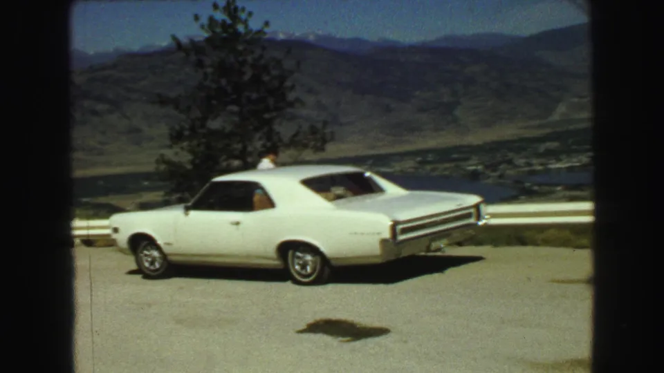 Home-movie frame of a white two-door car parked at a British Columbia scenic turnout, with lake water and mountains in the distance.