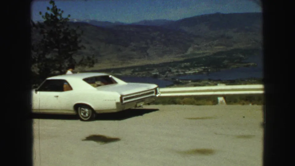 A white two-door car remains parked at the Vancouver-area overlook, seen with guardrail, distant lake, and mountain slopes on 8mm film.