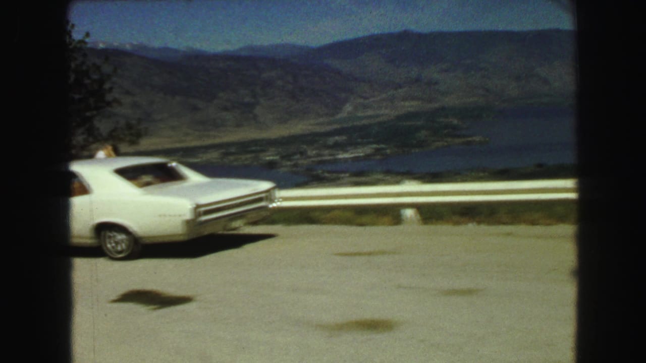 1968 8mm frame from Vancouver, British Columbia, showing a white sedan at a roadside overlook above a lake valley and distant hills.