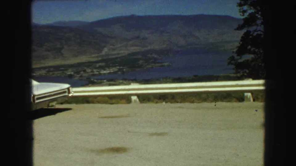 Elevated 1968 home-movie view near Vancouver, BC, with a guardrail, partial white car, blue lake, valley floor, and hills.