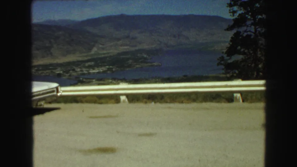 1968 8mm scenic overlook frame in Vancouver, British Columbia, with guardrail, lake water, valley settlement, hills, and roadside trees.