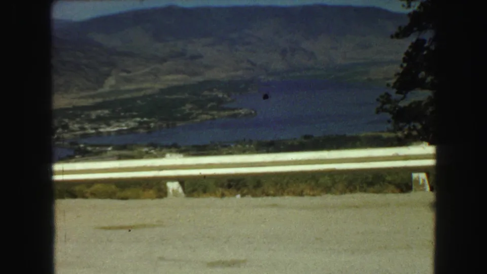Wide 1968 Vancouver-area home-movie view over a guardrail toward a lake, valley floor, wooded slopes, and distant mountains.