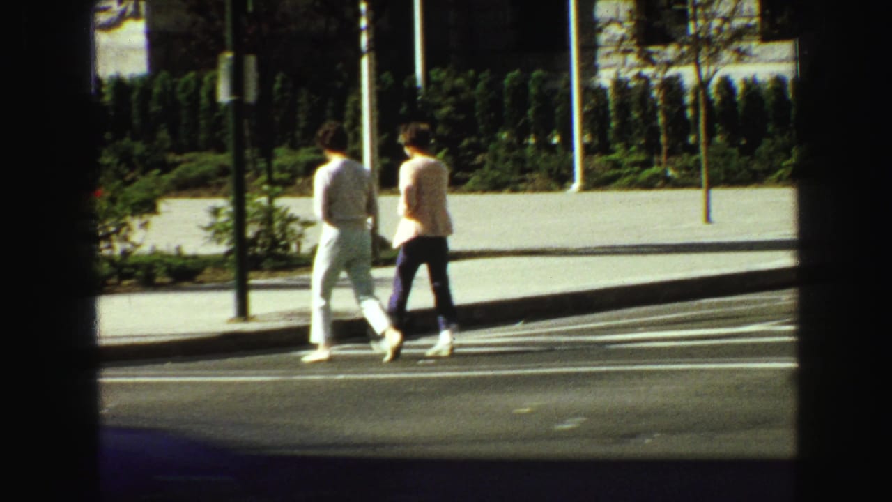 Two pedestrians walk along a sunlit sidewalk beside a city street.
