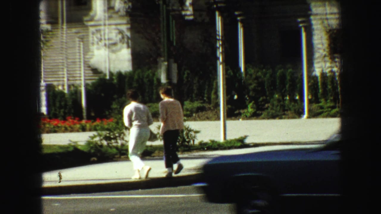 Two pedestrians continue along the sidewalk as a dark car passes in front.