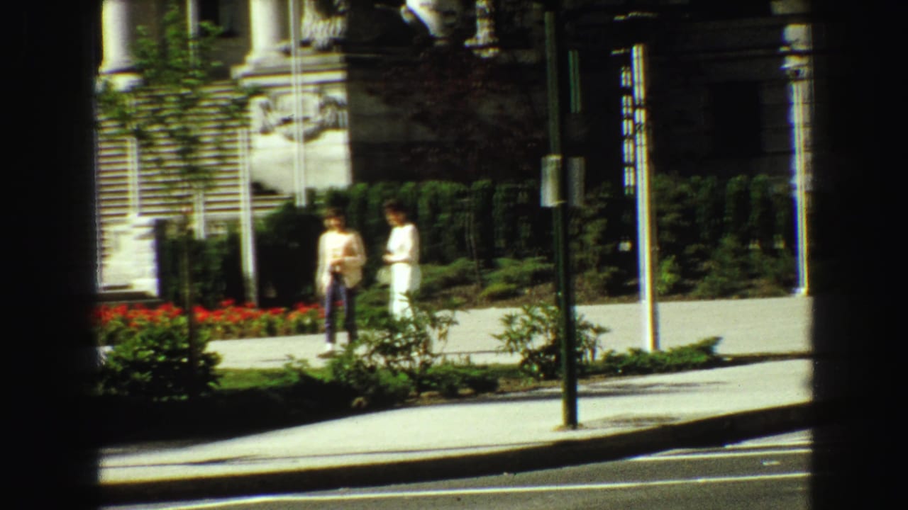 Two people in 1968 Vancouver appear near red flowerbeds and the steps of a large stone building on a sunny sidewalk.