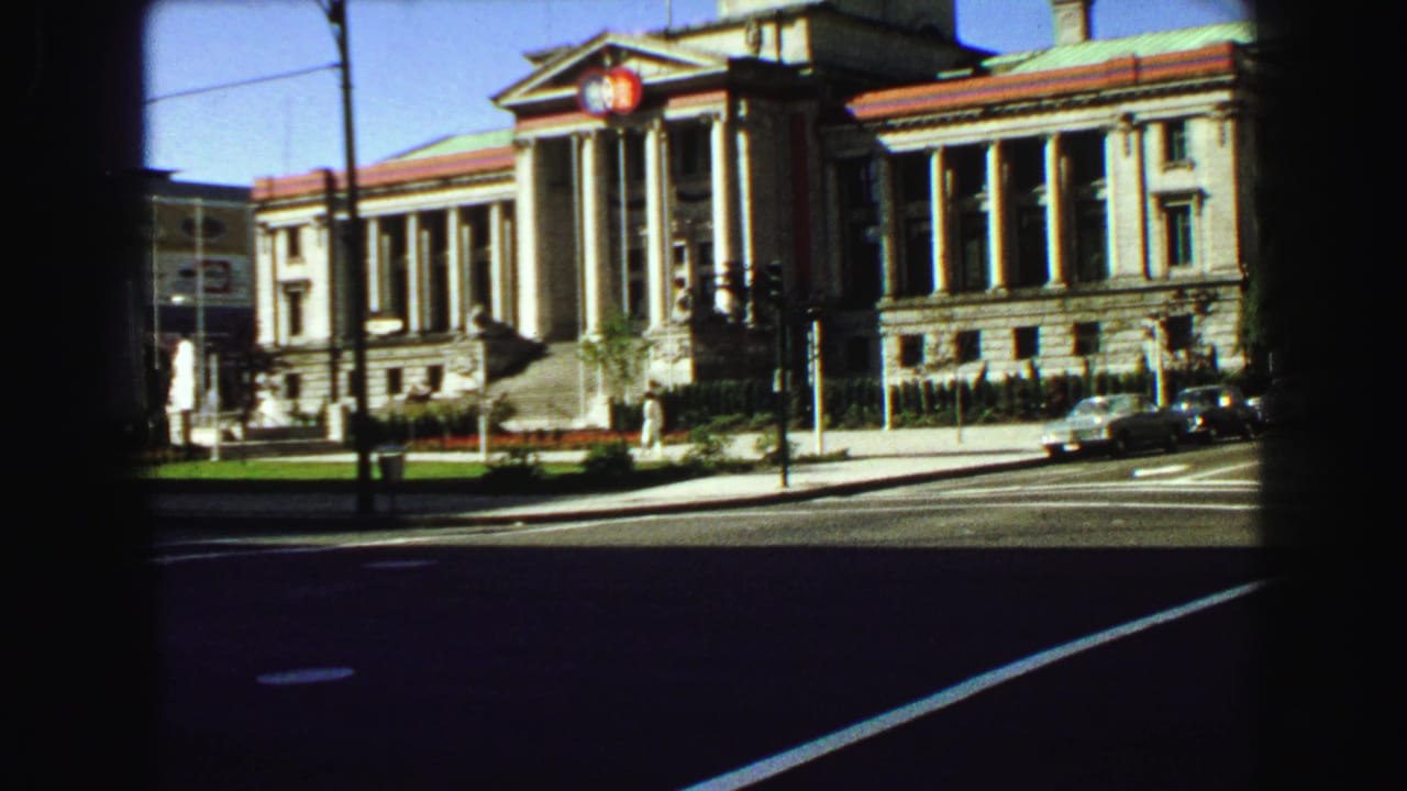 Columned stone building across a city street with period cars and a dark film border.