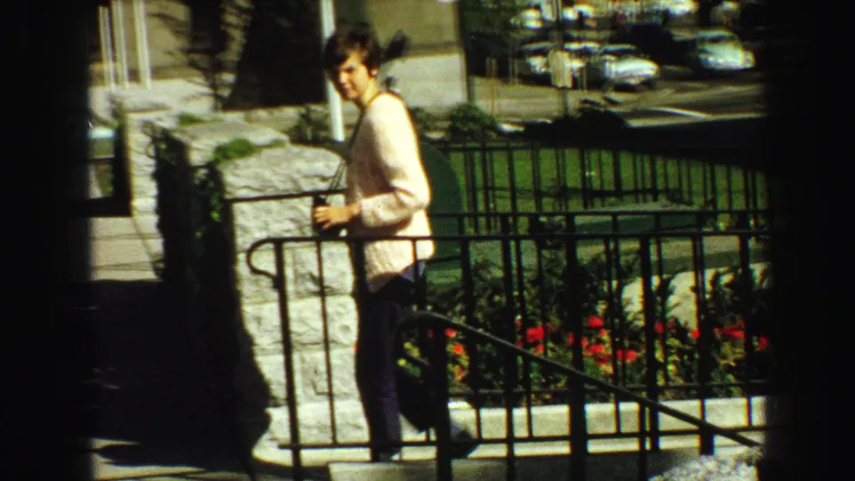 1968 Vancouver home-movie frame with a woman near a stone entrance, black railing, red flowers, and city street background.