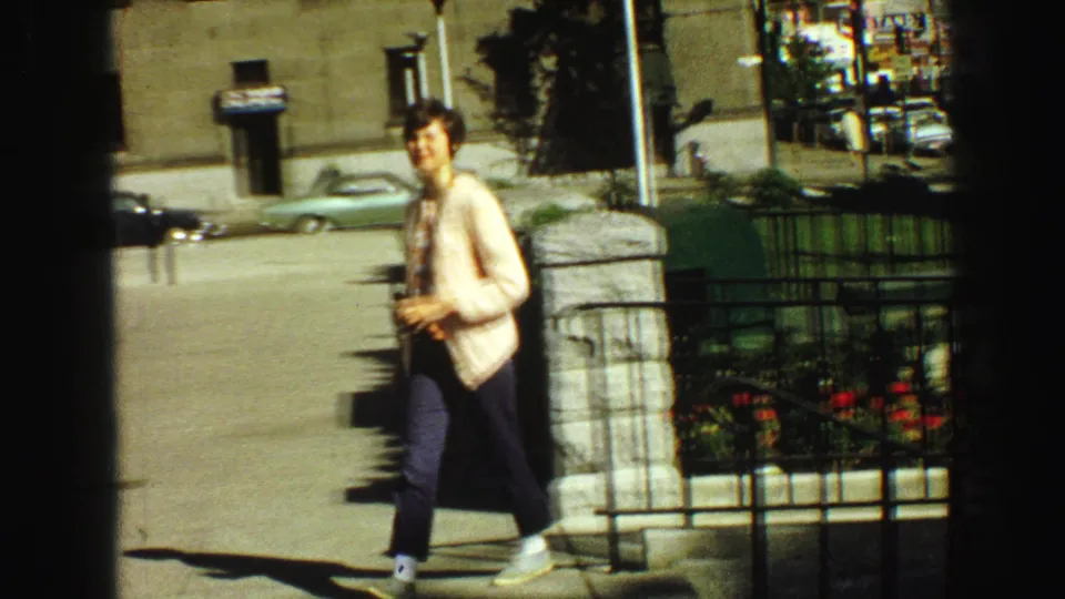 1968 Vancouver frame showing a smiling woman walking toward the camera beside a railing, stone post, sidewalk, and cars behind her.