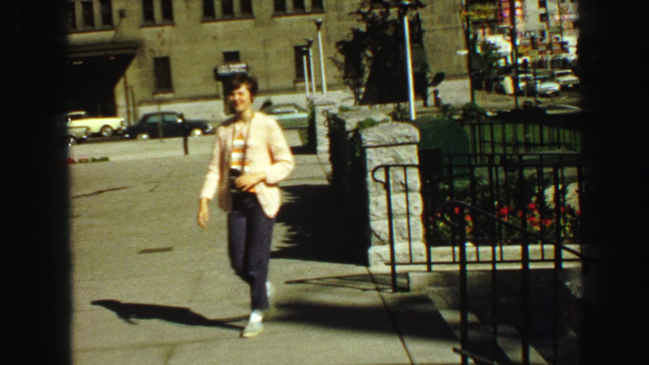 Young woman walks across a sunny paved square near a railing and stone building.