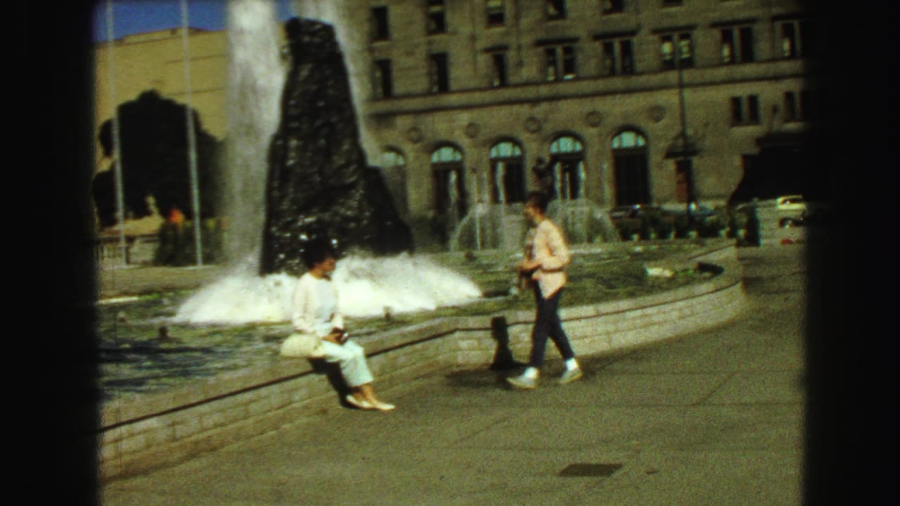 Young woman approaches another person seated on the edge of a large active fountain.