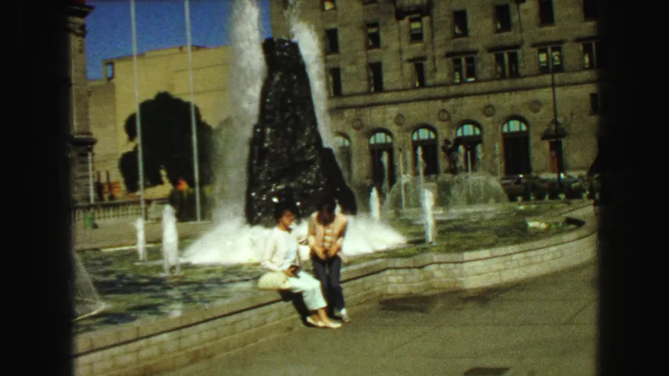 Two young women sit side by side at a fountain in 1968 Vancouver, with water jets and a stone building behind them.