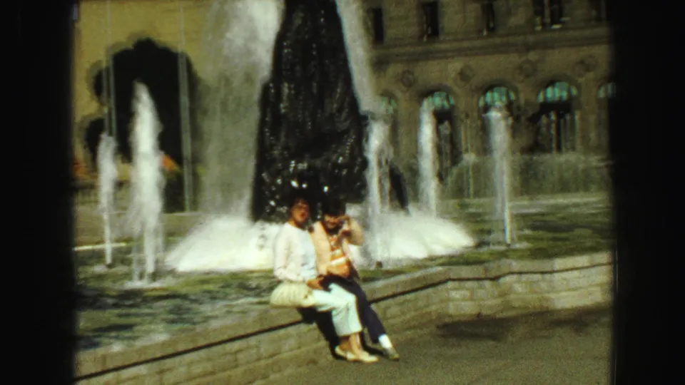 Two young women sit close together by the fountain, framed by water spray and a civic-looking stone facade.