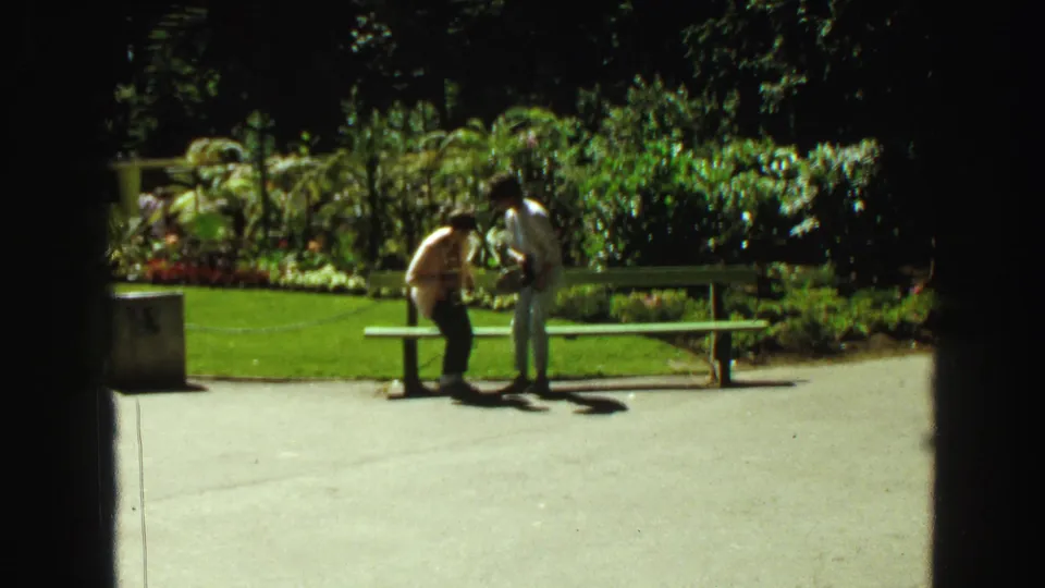 Two visitors stand beside a green bench in a sunny landscaped area, with garden beds and shrubs behind them.