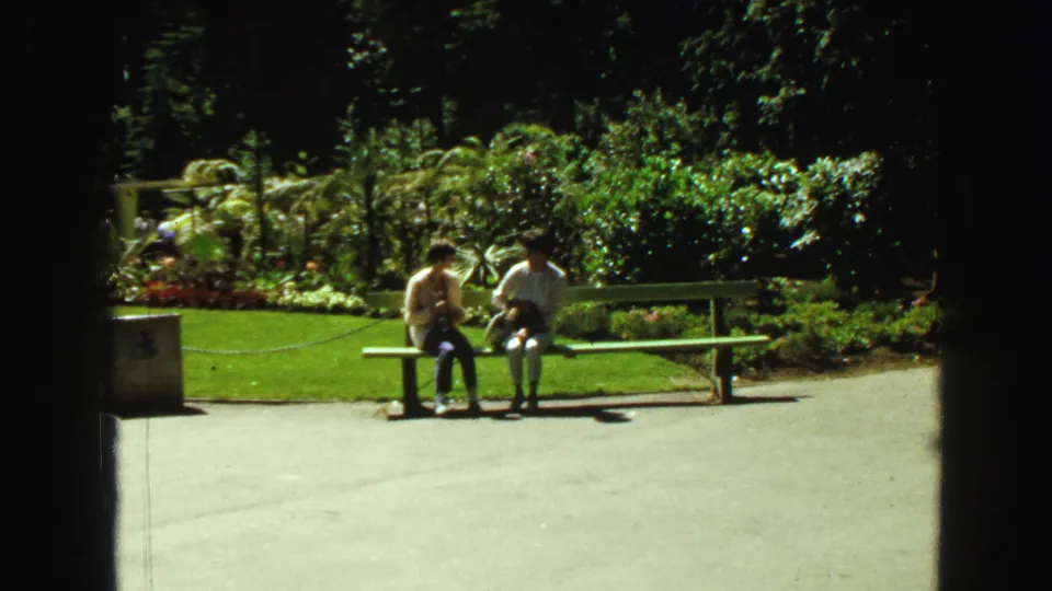 Two visitors sit on a green bench in the Vancouver zoo garden area, surrounded by bright plantings and paved walkways.