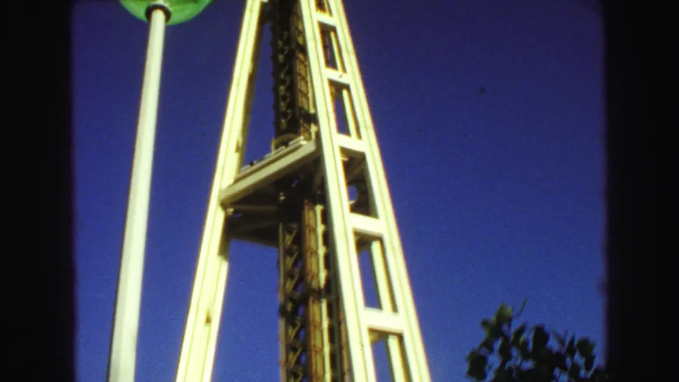 Low-angle 1968 view of the Space Needle elevator structure with a green globe lamp in the foreground and blue sky behind it.