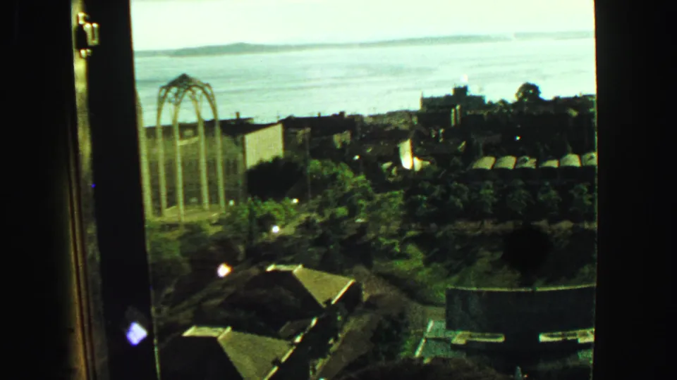 High observation-deck view over rooftops, trees, water, and distant shoreline, with window edges framing the city panorama.