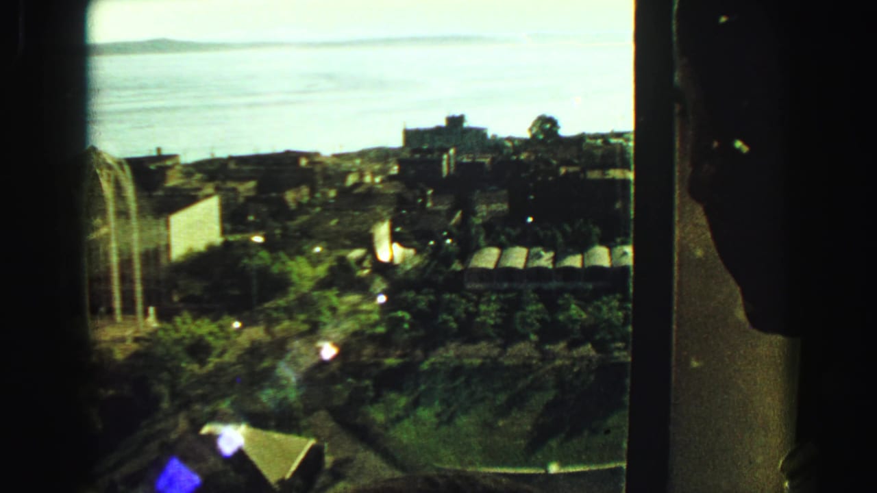 1968 Vancouver frame from behind a window, with harbor water beyond trees, rooftops, and a partially visible arched structure.