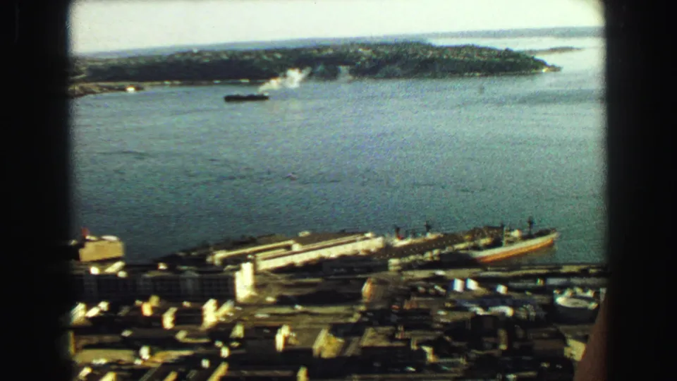 1968 Vancouver waterfront frame showing bay water, a docked ship, industrial roofs, pier structures, and distant shoreline.