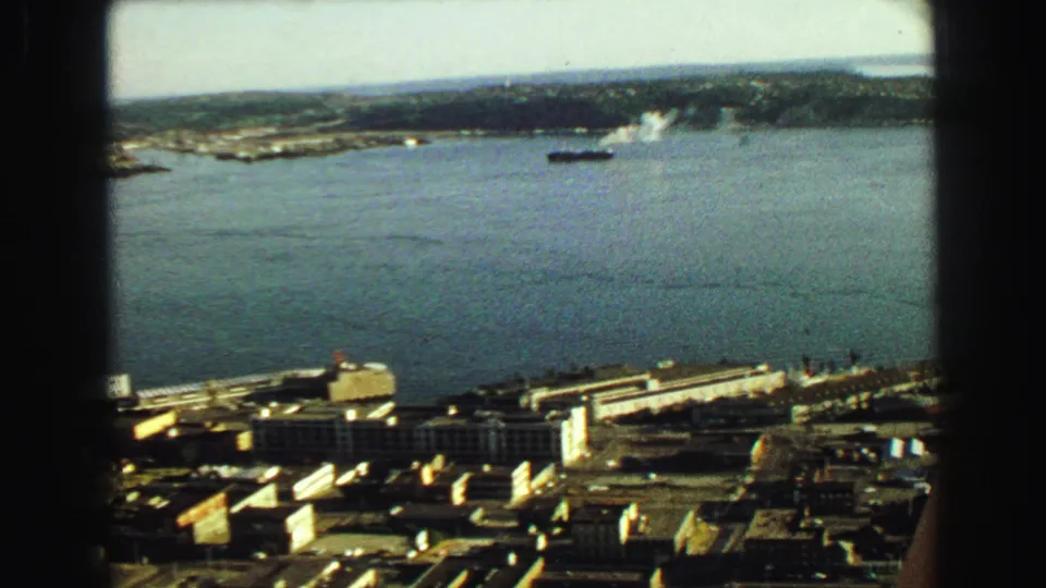 1968 Vancouver home-movie frame looking through a window at harbor docks, a large ship, open water, and distant land.